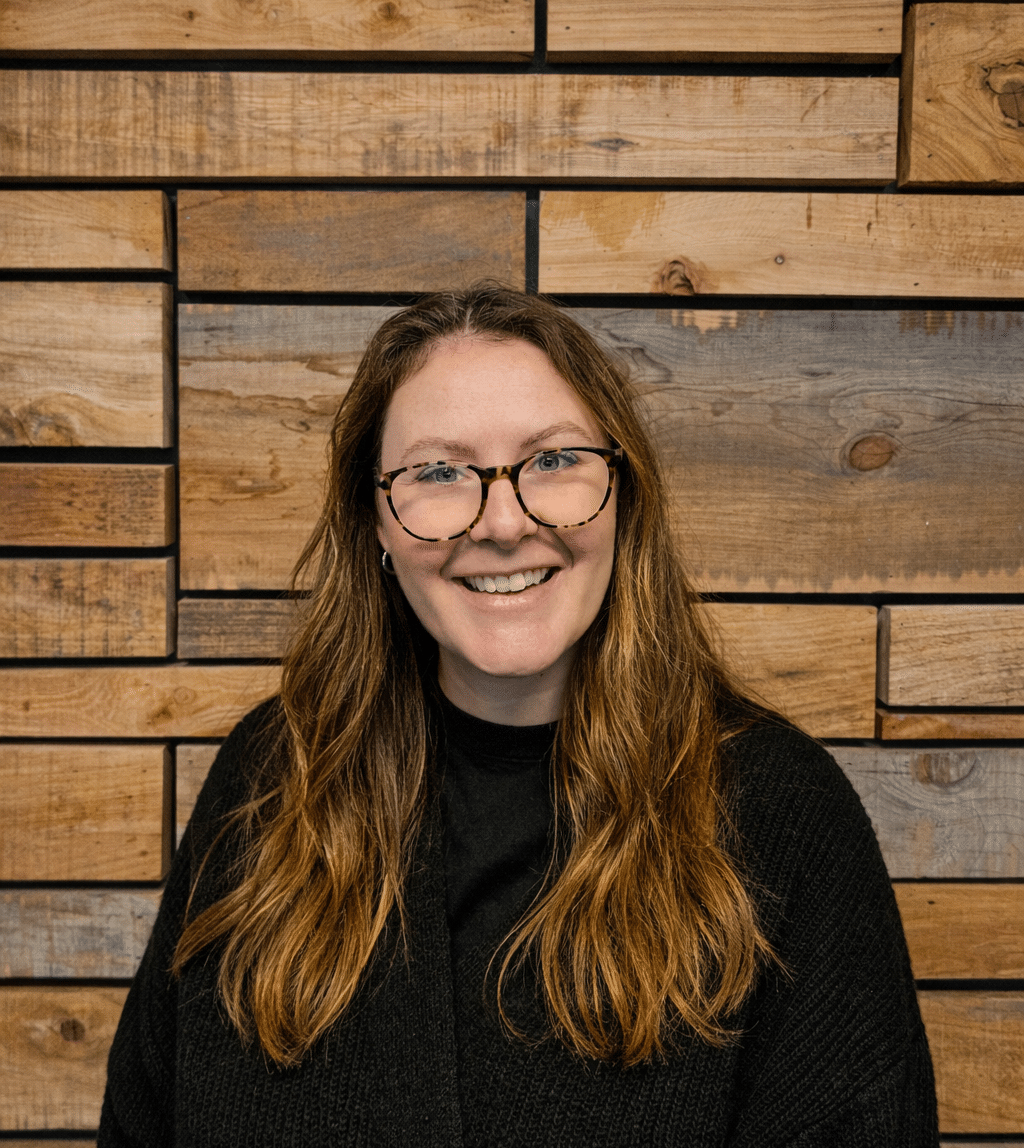 Smiling woman with long light brown hair and tortoiseshell glasses in front of a wooden paneled wall.