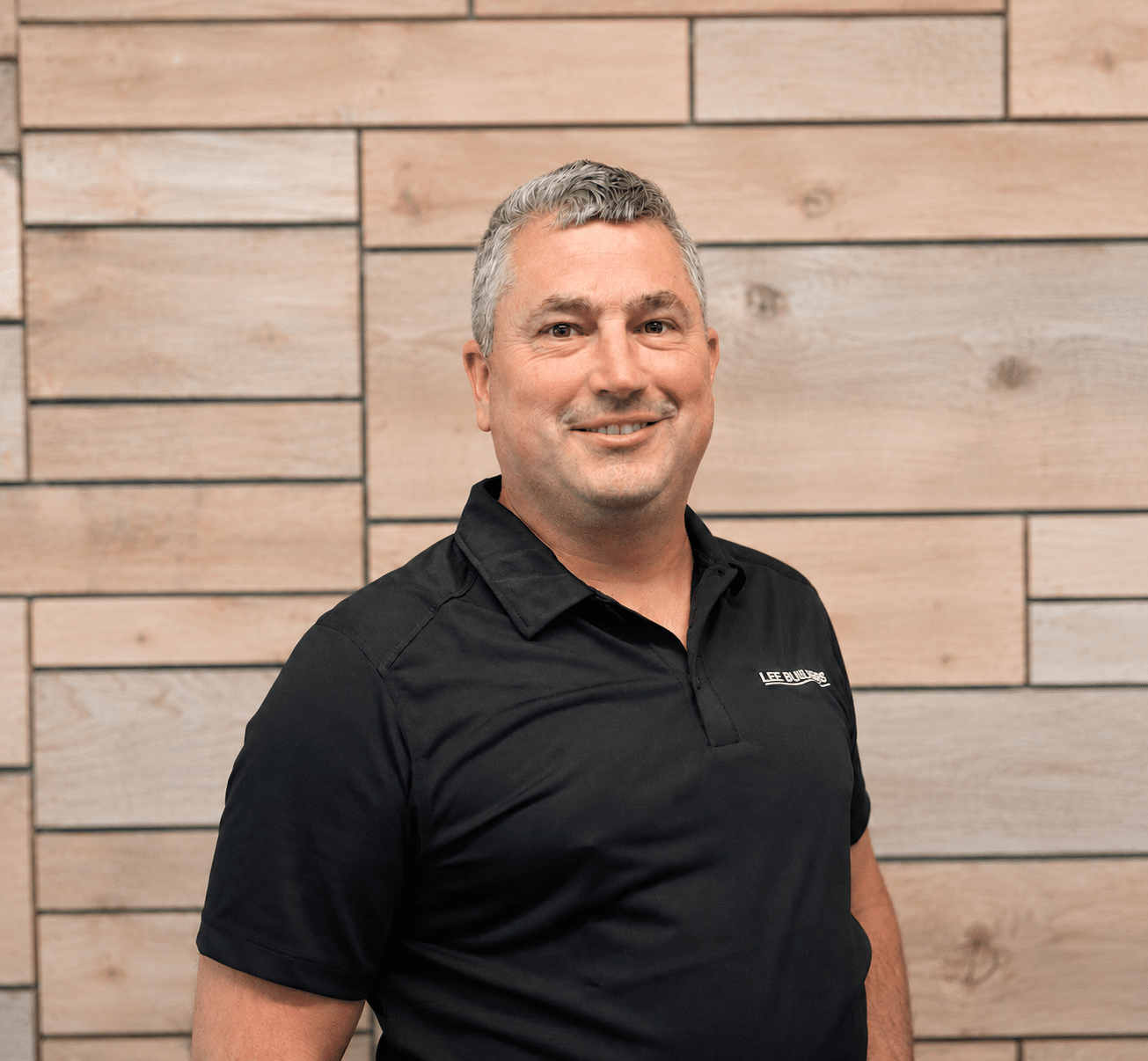 Portrait of a smiling man in a black polo shirt standing against a light wood panel wall.